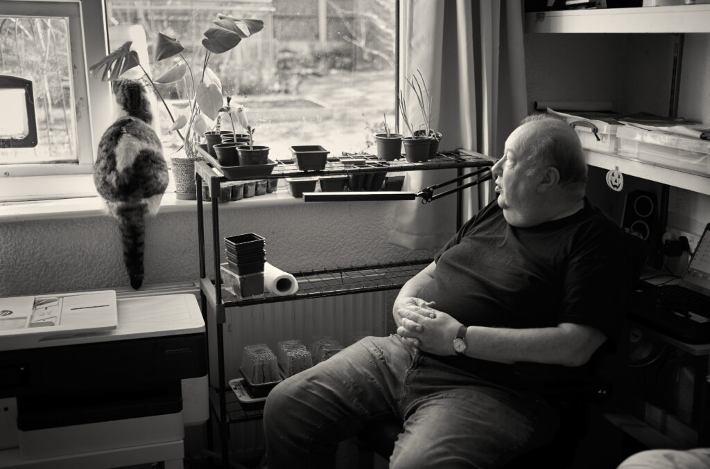 Artist Rob Lees in his bedroom with cat looking out into his garden. Shelves of seed trays in his room.