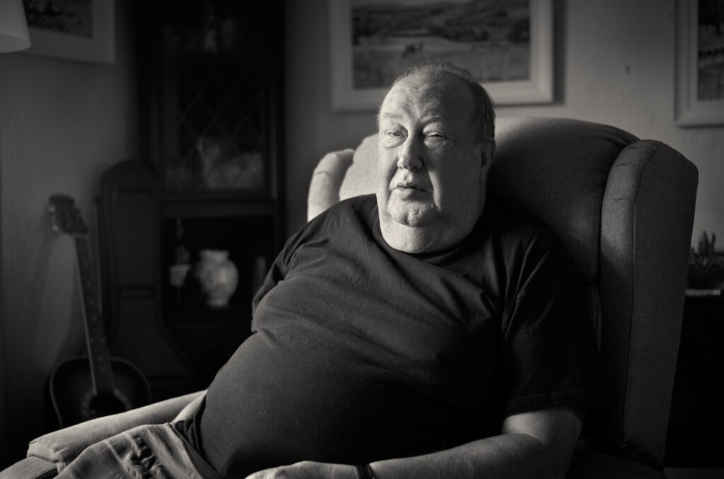 Artist Rob Lees, portrait of him sat in an armchair, his guitar visible behind him, lit by natural window light.